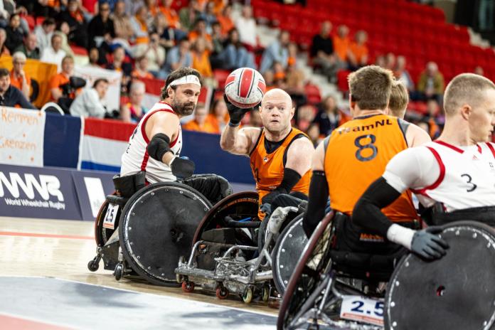 A male wheelchair rugby player wearing an orange jersey is throwing the ball during competition